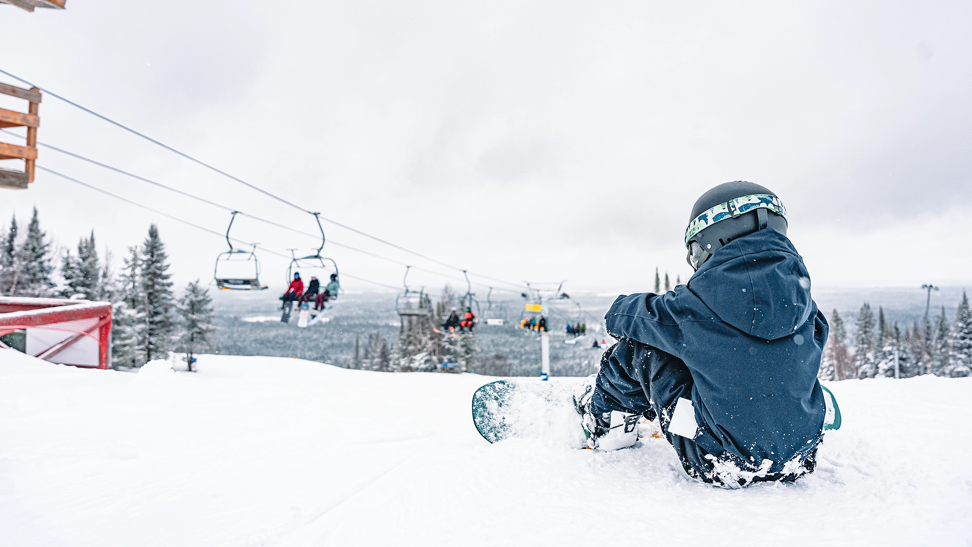 Un jeune planchiste est assis dans la neige au sommet de la montage avec les télésièges en arrière-plan